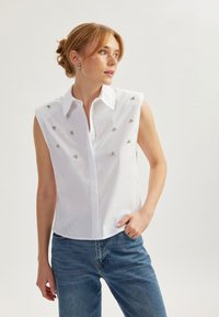 Blusa bianca senza maniche con colletto a punta, caratterizzata da embellishments argentati. Indossata con jeans in denim blu, dalla texture liscia e dalla vestibilità sartoriale.