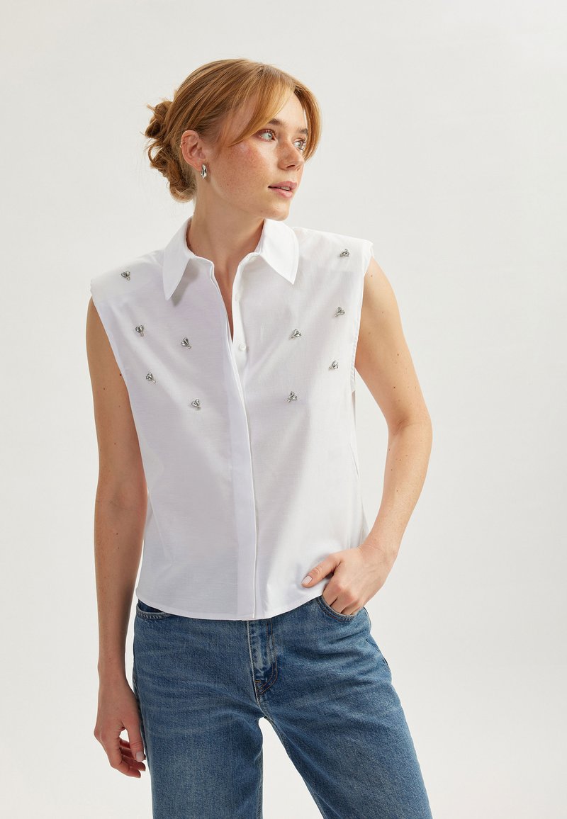 Blusa bianca senza maniche con colletto a punta, caratterizzata da embellishments argentati. Indossata con jeans in denim blu, dalla texture liscia e dalla vestibilità sartoriale.