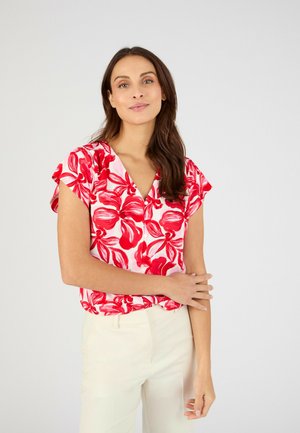 Mujer con cabello largo y castaño, vistiendo una blusa de manga corta con flores rojas y blancas y pantalones color crema, de pie con un brazo cruzado sobre el otro.