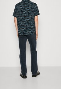 Chemise à manches courtes avec un fond noir et un motif de texte manuscrit bleu. Assortie à un jean foncé et des chaussures noires.