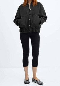 Mørkegrå fleece bomberjakke med store forlommer, trykknapper og ribbet kant, parret med sorte leggings og flade slip-on sko med nitter.