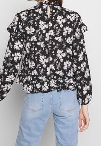 Mustakukkainen paita, jossa on valkoisia päivänkakkaroita, röyhelöhihoja, avaus selässä ja peplum-helma, valmistettu kevyestä kankaasta.