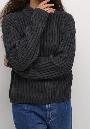Person wearing a dark gray ribbed knit sweater and blue jeans, with one hand near their face and the other arm crossed.