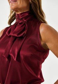 Bordeaux satin bluse med en slipsdetalje, uden ærmer, glat tekstur og en diskret lomme på venstre side.