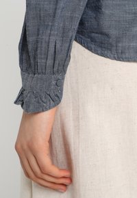 Blouse en lin bleu avec des manches froncées, présentant un poignet à volants et un détail de bouton. Le tissu a une finition texturée et légèrement brillante.