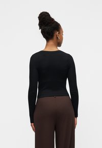 Γυναίκα με σκούρο δέρμα και μαλλιά σε κότσο, φορώντας στενή μαύρη μπλούζα μακρυμάνικη και φαρδιά καφέ παντελόνια, γυρισμένη ανάποδα με φόντο έναν απλό τοίχο.