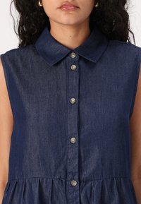 Sleeveless denim dress in dark blue with a collared neckline, button front, and gathered waist detail, featuring smooth fabric texture and subtle sheen.
