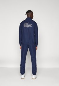 Marineblå træningsdragt lavet af glat stof, med et mønstret Lacoste-logo på ryggen, elastiske manchetter og sidelister på bukserne.