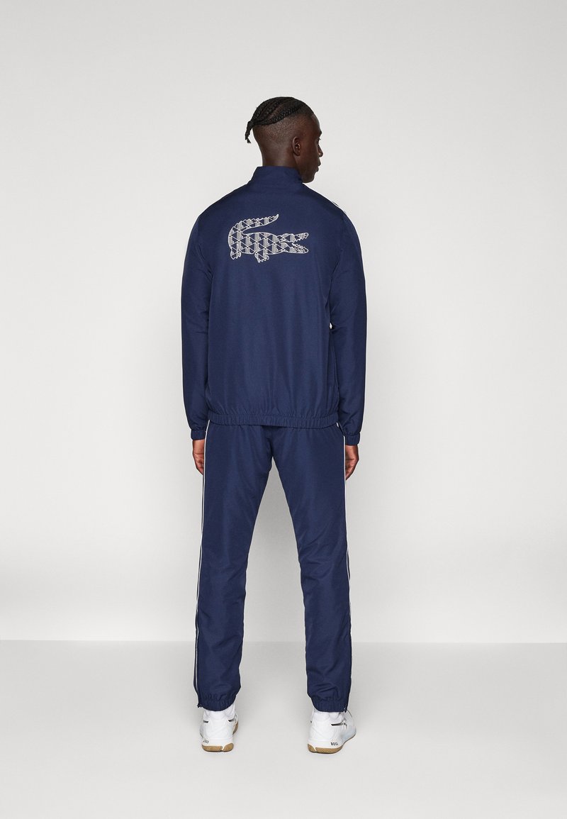 Marineblå træningsdragt lavet af glat stof, med et mønstret Lacoste-logo på ryggen, elastiske manchetter og sidelister på bukserne.