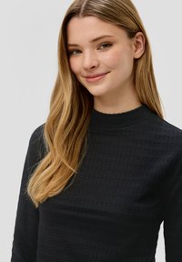 Crni teksturirani dukserica dugih rukava s visokim izrezom. Ima suptilan uzorak i glatku tkaninu. Model ima ravnu kosu.