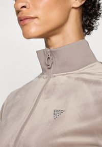 Light beige zip-up jacket with a high collar, made of soft material, featuring a triangular silver studded accent on the front.
