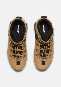 Bruna lädervandringsskor med en svart mesh-tunga, guldöglor och ljusa sulor. Har snörningsdesign och Timberland-logotyp.