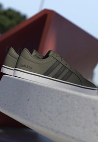 Par de zapatillas Adidas verde oliva con rayas negras y suelas blancas descansando sobre una superficie de piedra clara al aire libre.