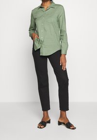 Chemise verte à manches longues avec boutons et poche poitrine, portée sur un pantalon noir ajusté et des talons ouverts noirs, avec une texture lisse.