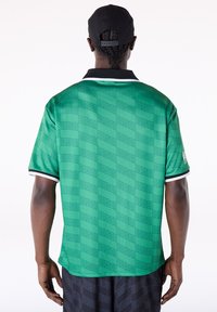Camiseta deportiva verde hecha de tela ligera, con un patrón repetido de "BOSTON", cuello y mangas negras, y mangas cortas.