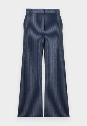 Mujer de pie con las manos en los bolsillos, vestida con un traje azul marino a rayas, pantalones anchos hasta el tobillo y zapatos Mary Jane de color rojo oscuro.