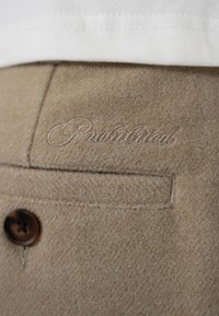 Poche d'un pantalon en laine beige avec un bouton marron et un texte brodé discret "Produced" au-dessus de la poche, sous le bord d'un vêtement blanc.