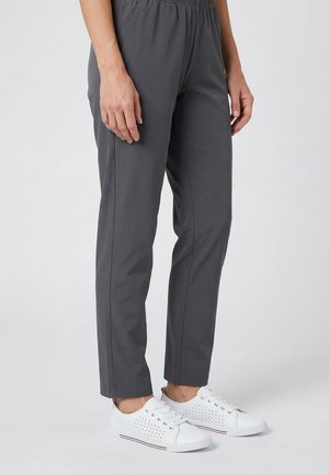 Vrouw met donkere grijze slim-fit broek en witte geperforeerde vetersneakers, staand tegen een effen lichte achtergrond.