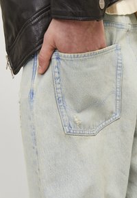 Lys blå denimjeans med en avslappet passform, med en baklomme med et lite hull og et synlig søm mønster. Hånd som hviler i lommen.