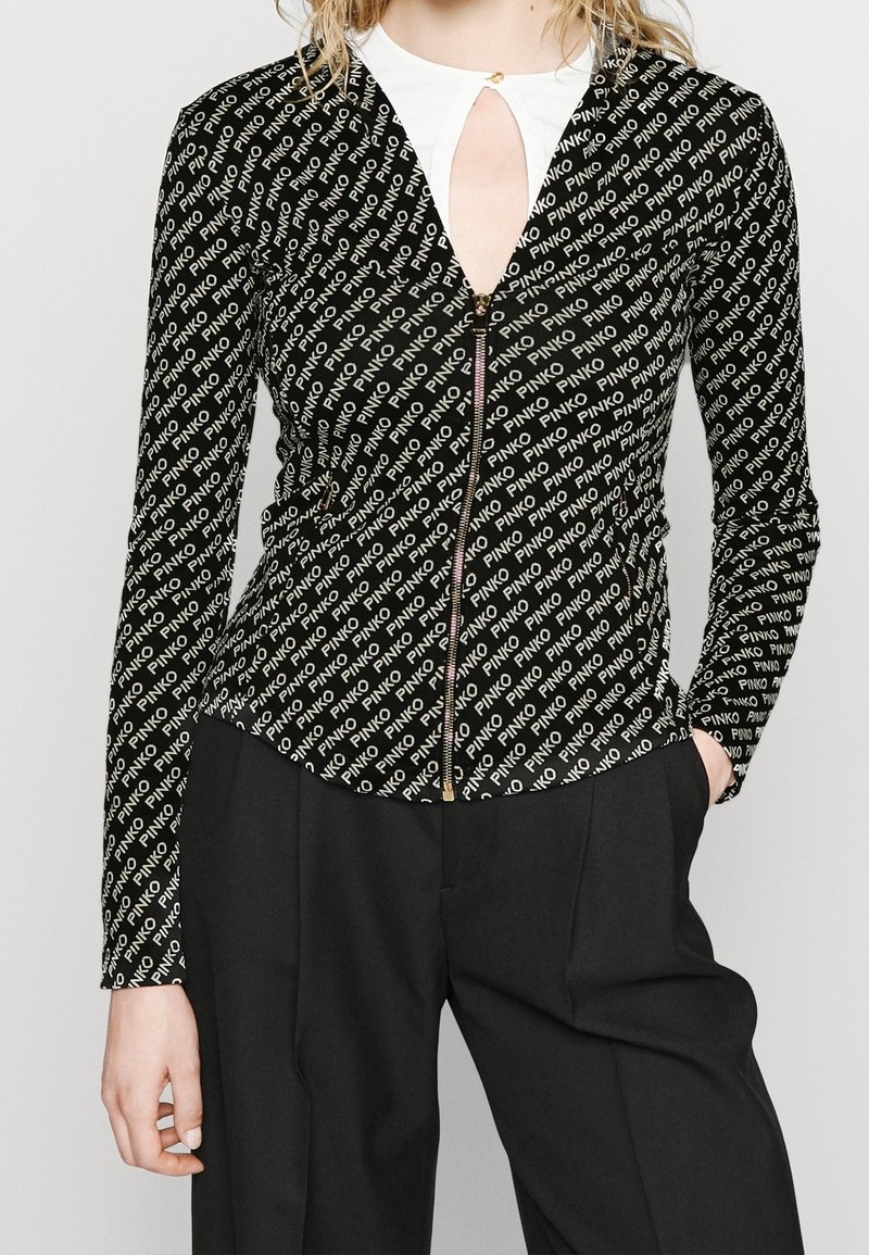 Femme portant une veste noire zippée avec un motif de texte blanc "PINKO", un haut blanc avec une encolure en trou de serrure et un pantalon noir, les mains dans les poches.