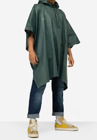 Grøn poncho med hætte, lavet af letvægts, blankt materiale. Har snoretræk-detaljer og et løst, kappe-lignende design. Bæres over blå jeans.