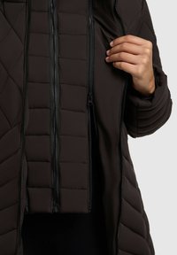 Brun, quiltet pufferjakke med lynlås foran. Indvendigt har den glat stof. Detaljerne inkluderer lodrette syninger og lynlås-lommer.