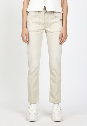 Frau trägt cremeweiße, hoch taillierte Jeans mit geraden Beinen und vorderen aufgesetzten Taschen sowie weiße Sneakers, steht vor einem einfarbigen Hintergrund.