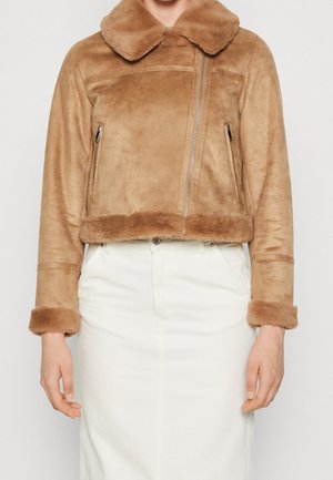 Vrouw die een korte beige nepbontjas met kraag, ritssluiting en witte rok draagt tegen een effen achtergrond.