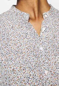 Lähikuva valgest bluusist, millel on väiksed roosad lilled ja mustad täpid, mandariinikaelus ja valged nööbid.