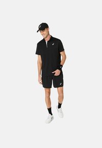 Uomo che indossa abbigliamento sportivo Asics nero, compresi cappellino, polo, pantaloni corti, fasciatura per il polso, calze nere e sneakers bianche, sorridendo e camminando.
