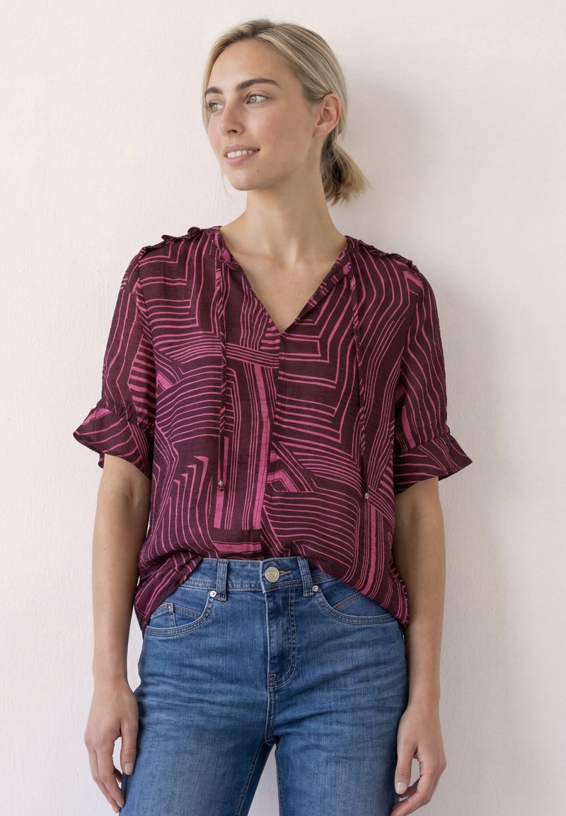 Femme portant un chemisier bordeaux avec des lignes géométriques roses et un jean bleu, debout contre un mur clair, regardant sur le côté avec un léger sourire.