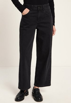 Person wearing black wide-leg jeans, black loafers, and a black ribbed long-sleeve top with one hand in pocket.