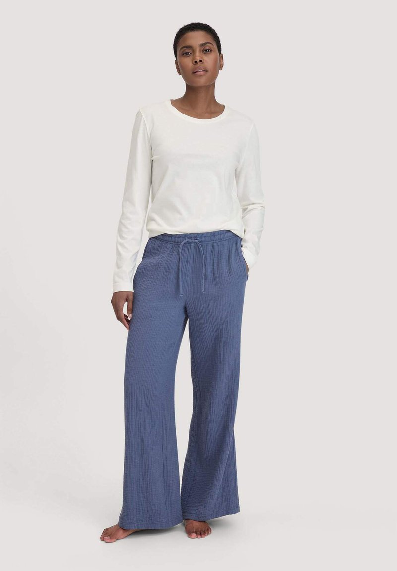 Eine Frau steht barfuß und trägt ein langärmliges weißes Oberteil sowie lockere blaue Jogginghosen vor einem einfachen hellen Hintergrund.