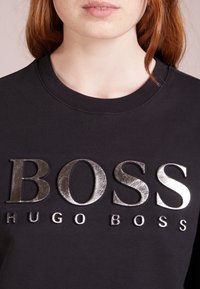 Svart sweatshirt med metallisk silver "BOSS" och "HUGO BOSS" logotyper. Klassisk rund halsringning och mjuk textur, med synliga sömnadsdetaljer.