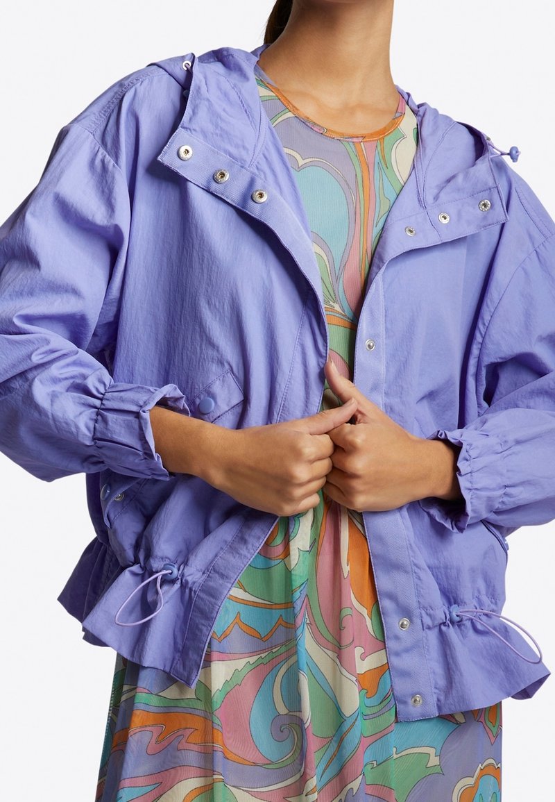 Světle fialová bunda s kapucí, elastickými manžetami a stahovací lem. Přes ni je vrstvené barevné šaty s vzory, které obsahují víry v pastelových barvách.