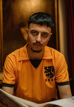 Man met kort zwart haar en sikje, in een oranje KNVB-shirt, kijkt naar de camera, zittend binnen met een wazige achtergrond.