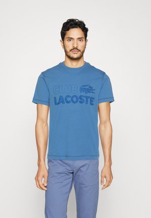 T-shirt en coton bleu avec le logo "CLUB LACOSTE" en bleu plus foncé, col rond, manches courtes et design simple à ourlet plat.