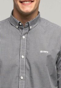 Koszula w kratę w czarno-białym kolorze, z klasycznym kołnierzykiem, białymi guzikami i małym logo na lewej piersi.
