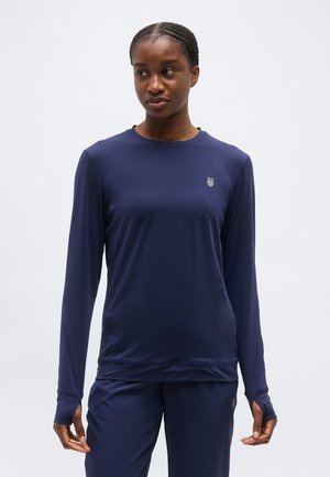 Moteris su pintinėmis plaukais, apsirengusi tamsiai mėlyna ilgomis rankovėmis palaidine ir derančiomis kelnėmis, stovinti ir žvelgianti šiek tiek į šoną.