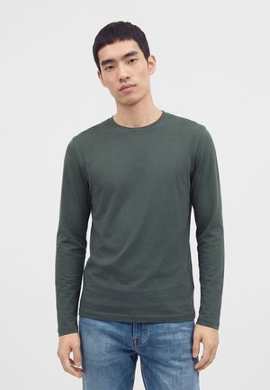 Ung mand med kort sort hår iført en mørkegrøn langærmet skjorte og blå jeans, stående mod en ensfarvet lys baggrund.