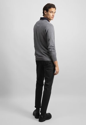 Jeune homme debout, portant un pull gris par-dessus une chemise noire à col, un pantalon noir et des chaussures noires, regardant par-dessus son épaule.