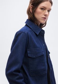 Νεαρή γυναίκα με ανοιχτόχρωμο δέρμα και καστανά μαλλιά, φορώντας μια σκούρα μπλε τζιν ζακέτα με μεγάλες μπροστινές τσέπες, κοιτάζοντας ελαφρώς προς το πλάι.