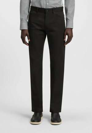 Zwarte broek met een rechte snit, gemaakt van een gladde stof. Voorzien van zijzakken, een knoopsluiting en gecombineerd met zwarte schoenen.