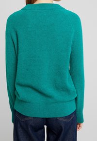 Türkiser Pullover aus weichem Strickmaterial mit Rundhalsausschnitt und geripptem Saum. Kombiniert mit einer dunklen Jeans, Blick von hinten.