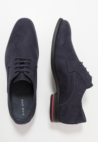 Chaussures habillées en daim bleu marine avec une texture lisse, un laçage traditionnel, et une semelle noire accentuée de rayures rouges et blanches au niveau du talon.