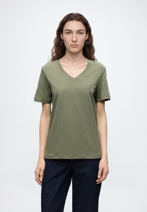 Jeune personne aux longs cheveux bouclés portant un t-shirt vert olive à col en V et un jean foncé, debout devant un fond blanc uni.