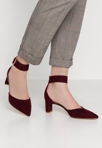 Burgundy mockask komfortskor med hög klack, spetsig tå och ankelsnodd med ett runt detaljer. Klacken mäter cirka 6,5 cm.