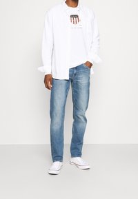 Camisa blanca de botones sobre una camiseta gráfica, jeans azules y zapatillas blancas. Ajuste casual; la camisa tiene mangas largas y la camiseta presenta un logo.