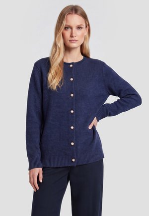 Kvinde med langt blond hår iført en navyblå knap-cardigan og mørke bukser står med den ene hånd på hoften.