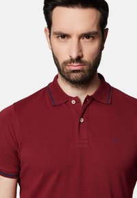 Burgundowa koszulka polo o strukturalnym wykończeniu, z dwoma guzikami i kontrastowym granatowym wykończeniem kołnierza. Posiada małe logo na klatce piersiowej.